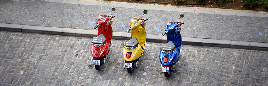 En röd, en gul och en blå vespa står parkerade vid en trottoarkant