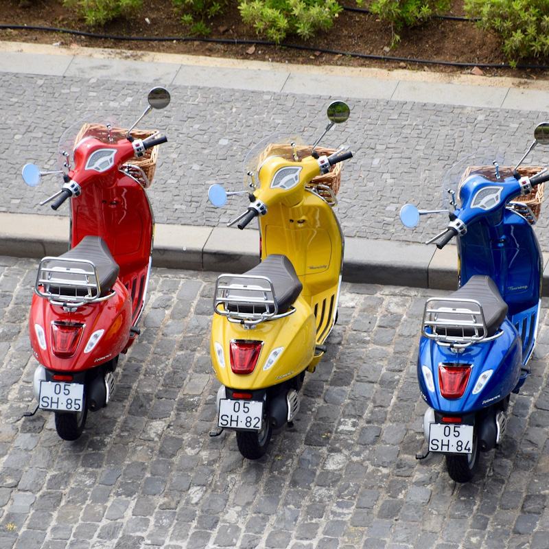 En röd, en gul och en blå vespa står parkerade mot en trottoarkant